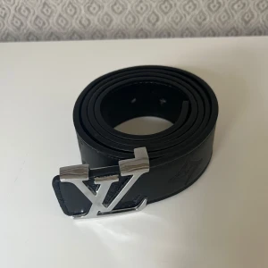 Svart bälte i skinn - Snyggt svart bälte i skinn med en stilren silverfärgad logga som spänne. Bältet har en klassisk design och diskret präglad logga på lädret. Perfekt accessoar för att lyfta din outfit.