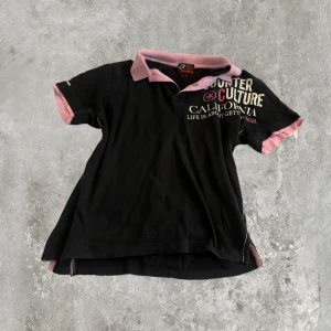 Svart piké  - Cool svart t-shirt från Counter Culture med massor av tryck både fram och bak. Har rosa krage och rosa detaljer vid ärmslut. T-shirten har korta ärmar och en klassisk pikékrage. Materialet är mjuk bomull, perfekt för en avslappnad streetstil.