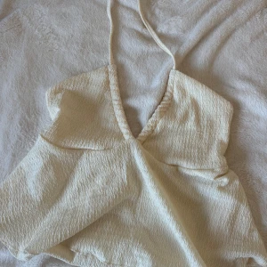 Krämvit halterneck linnetopp - Säljer en krämvit linnetopp med halterneck och djup v-ringning. Toppen har en vågig struktur i tyget som ger en cool effekt och är ärmlös med tunna knytband i nacken. Perfekt för varma dagar och somriga outfits. Är inte alls säker på storlek då den inte har någon lapp men den är xs eller S