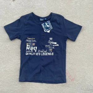 Mörkblå t-shirt från Xux med tryck - Snygg mörkblå t-shirt från Xux med coolt tryck och texten 'Qualified Legends' framtill. T-shirten har tre knappar vid halsen och är gjord i mjuk bomull. Perfekt för dig som gillar sportig och avslappnad stil.