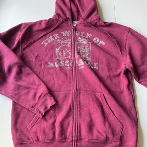 Vinröd hoodie med dragkedja från One of one - Säljer en vinröd hoodie från One of one med texten 'The Wolf of Mosebacke'. Tröjan har huva med snörning, fickor framtill och hel dragkedja. Perfekt för dig som gillar streetwear och vill ha något unikt.