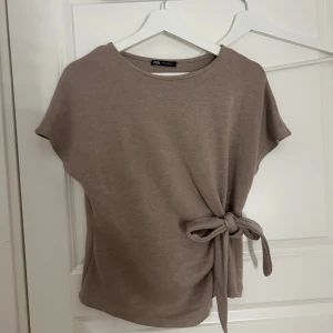 Beige topp med knyt från Zara - Säljer en beige topp från Zara med korta ärmar och snygg knytning i sidan. Toppen har rund halsringning och är i mjukt material som känns skönt mot huden. Perfekt för en stilren och avslappnad look.