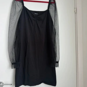 Svart klänning med meshärmar BIK BOK - Svart kort klänning från BIK BOK med transparenta meshärmar och smala axelband. Klänningen har en enkel, rak passform och diskreta rynkdetaljer vid halsen. Perfekt för dig som gillar trendiga detaljer och stilren look.