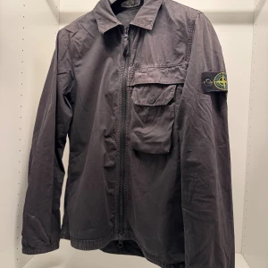Svart overshirt från Stone Island - Svart overshirt från Stone Island med klassisk krage, dragkedja framtill och en stor bröstficka med knapp. Ikonisk logotyp på vänster ärm. Jackan är tillverkad i bomull och har långa ärmar med elastiska muddar. Perfekt för lager-på-lager och streetwear.