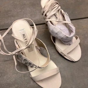 Klackar från NLY Shoes - Eleganta beige klackar med klack från NLY Shoes. Skorna har en stilren design med knytning runt ankeln och dekorativa stenar på remmen. Helt nya. 
