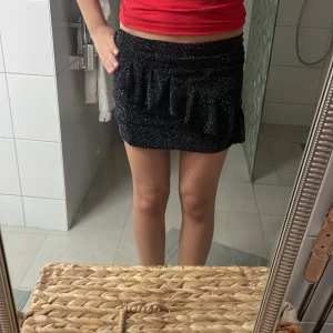 Glittrig svart minikjol med volang - Säljer en svart glittrig minikjol med volangdetaljer framtill. Kjolen har en tight passform och är perfekt för dig som vill sticka ut. Materialet är stretchigt och skimrar snyggt i ljuset. Supersnygg att styla med en enkel topp eller crop.