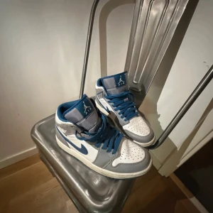 Nike Air Jordan 1 Mid Blå/Vit - Säljer ett par Nike Air Jordan 1 Mid i blått, vitt och grått. Skorna har klassisk hög siluett, blå snörning och vit perforerad tå. Swoosh-logga i blått på sidan och ikonisk Air Jordan-vinge på hälen. Ovandel i skinn och syntet, yttersula i blått gummi. Storlek us 8
