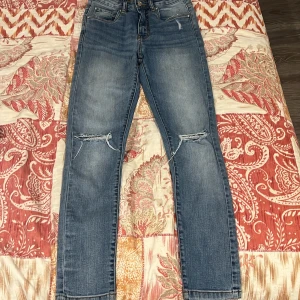 Blå jeans från KappAhl, strl 152 - Snygga blå jeans från KappAhl i storlek 152. Modellen har raka ben, slitna detaljer vid knäna och klassisk femficksdesign. Jeansen är tillverkade i hållbar denim och har normal passform med knapp och dragkedja framtill.