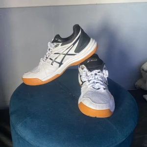 Asics Upcourt 4 vita sneakers - Säljer ett par Asics Upcourt 4 sneakers i vitt med svarta detaljer och orange sula. Skorna har snörning, meshpaneler för andning och klassisk Asics-logga på sidan. Perfekta för inomhussport eller träning. Snygg och sportig design som sticker ut.