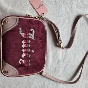 Vinröd axelväska från Juicy Couture - Juicy Couture axelväska i vinröd mocka med ljusrosa skinn-detaljer och justerbar axelrem. Väskan har broderad Juicy-logga i vitt och rosa på framsidan och guldfärgade metalldetaljer. Perfekt storlek för mobil, plånbok och småsaker.