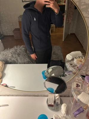 Mörkblå hoodie från Polo Ralph Lauren - Säljer en mörkblå hoodie från Polo Ralph Lauren med vit broderad logga på bröstet. Tröjan har huva med snörning, dragkedja framtill och två fickor. Materialet är mjuk bomull och passformen är normal. Perfekt för chill dagar eller när du vill vara extra comfy.