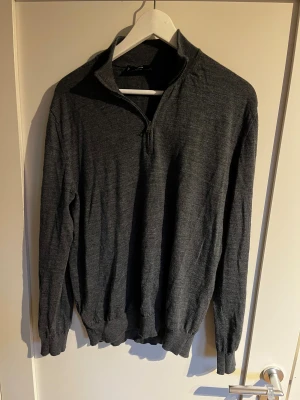 Mörkgrå merinoullströja från Mango - Snygg mörkgrå långärmad tröja från MNG Essentials i mjuk merinoull. Tröjan har en halv dragkedja vid kragen och ribbade muddar vid ärmslut och nederkant. Perfekt för dig som gillar stilren och enkel design med lite sportig touch.