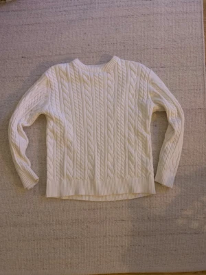 Vit kabelstickad tröja Kappahl - Säljer en vit kabelstickad tröja från Kappahl i storlek 146/152. Tröjan har ett klassiskt flätmönster, rund hals och ribbade muddar vid ärmslut och nederkant. Perfekt för dig som gillar en clean och mysig stil under kyliga dagar.