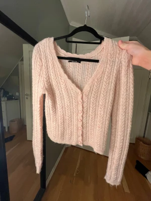 Ljusrosa stickad kofta från Bikbok - Supermysig ljusrosa stickad kofta från BikBok med snyggt flätmönster och v-ringning. Koftan har långa ärmar och knappar framtill, perfekt för att ge din outfit en soft och chill vibe. Passar dig som gillar en feminin och cozy stil.