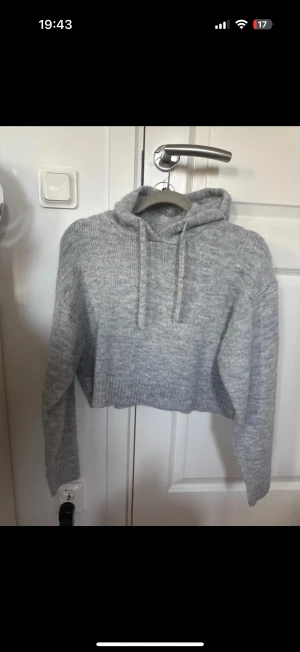 Grå croppad stickad hoodie - Säljer en grå croppad stickad hoodie med huva och snörning. Tröjan har långärmade ribbade ärmslut och en mjuk, mysig känsla. Perfekt för dig som gillar en chill och trendig look med lite oversized vibe.