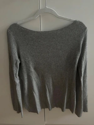 Grå ribbad tröja med omlott i ryggen - Säljer en grå ribbad omlottopp med lång ärm och bred halsringning. Toppen har en snygg omlott-detalj baktill. Perfekt för dig som gillar stilrena basplagg med en twist