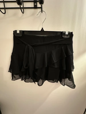 Svart volangkjol med knytband - Snygg svart kortkjol med flera lager volanger och ett dekorativt knytband i midjan. Kjolen är gjord i ett lätt och luftigt syntetmaterial som ger en skön känsla och snygg rörelse. Perfekt för dig som gillar en feminin och trendig stil.