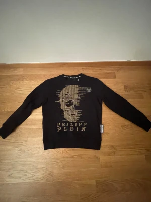 Svart tröja med guldtryck - Svart långärmad tröja med guldiga nitar som bildar en döskalle och logotyp på framsidan. Baksidan har stort PHILIPP PLEIN-tryck i guld. Tröjan har rund hals och ribbade muddar. Materialet är mjuk bomull med exklusiva detaljer.