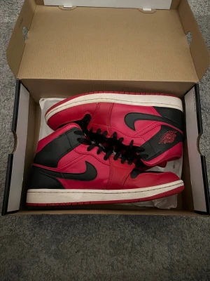 Nike Air Jordan 1 röd/svart sneakers - Säljer ett par Nike Air Jordan 1 sneakers i klassisk röd och svart färg med vita sulor. Skorna har högt skaft, svart snörning och ikoniska Jordan Wings-loggan på sidan. Materialet är skinn och syntet, med rund tå och platt sula. Perfekt för dig som gillar streetstyle och vill sticka ut.