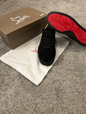 Christian Louboutin svarta sneakers - Snygga svarta sneakers från Christian Louboutin i mocka med klassisk röd sula och svarta snören. Låg modell med rund tå och diskret branding på insidan. Perfekta för dig som vill ha en lyxig och stilren look med en ikonisk touch.