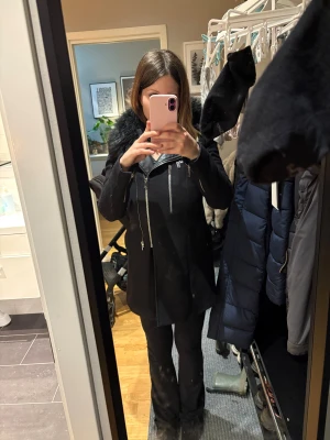 Svart kappa med fuskpäls och dragkedjor - Säljer en svart kappa med snygga silvriga dragkedjor och fuskpäls vid kragen. Jackan har lång ärm, rak passform och flera dragkedjedetaljer framtill. Perfekt för dig som vill ha en stilren och cool look under kallare månader.