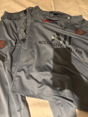 Blå PSG fotbollströja från Nike - Snygg blå fotbollströja från Nike med PSG-logga på bröstet och Accor Live Limitless-tryck. Tröjan har korta ärmar och en röd Nike-swoosh. Materialet är lätt och andas, perfekt för träning eller match. Ikonisk design med diskreta detaljer.