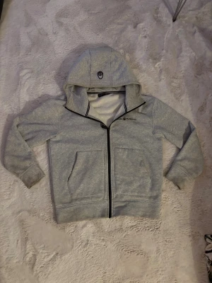 Grå hoodie från Sail Racing med dragkedja - Snygg grå hoodie från Sail Racing med svart dragkedja och två stora fickor framtill. Tröjan har huva med logga och långärmade ärmar. Perfekt för chill dagar och har en clean look som funkar till jeans eller joggers.