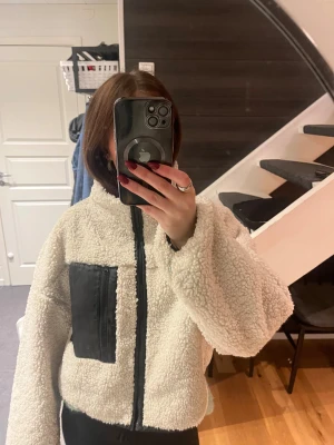Vit teddyjacka med svart ficka - Säljer en fluffig vit teddyjacka med svart dragkedja och svart ficka på bröstet. Jackan har hög krage och är långärmad, croppad i modellen. Jackan är köpt på Gina Tricot och är välanvänd. Den är i storlek M men passar mig som normalt är S/XS