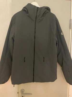 Grå vindjacka från sail racing - Snygg grå vindjacka från sail racing  med huva och hel dragkedja framtill. Jackan har två fickor med dragkedja, diskret logga på ärmen och justerbar nederkant. Perfekt för dig som gillar stilren och funktionell design.