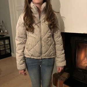 Beige quiltad pufferjacka med dragkedja - Säljer en beige pufferjacka med quiltat mönster och hög krage. Jackan har dragkedja framtill och en loose passform som ger en chill vibe. Perfekt för kyliga dagar och enkel att matcha med jeans eller hoodies.