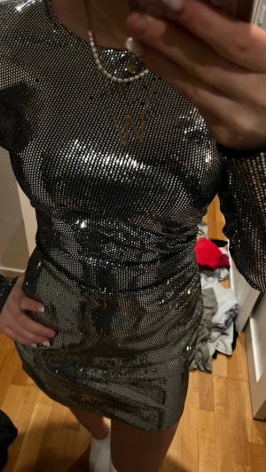 Glittrig silver kortklänning - Säljer en glittrig silverfärgad kortklänning med lång ärm och rund hals. Klänningen har en tight passform och är täckt av små paljetter som ger en riktigt snygg och festlig look. Perfekt för dig som vill sticka ut och glänsa på nästa event! Några få paljetter har lossnat som man kan se på bilden! Annrs i topp skick
