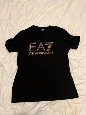 Svart EA7 Emporio Armani t-shirt XL - Svart t-shirt från EA7 Emporio Armani i storlek XL. Snyggt tryck i guld på bröstet med EA7-loggan. Klassisk rund hals och korta ärmar. Mjuk och skön bomullskvalitet som känns riktigt nice mot huden.
