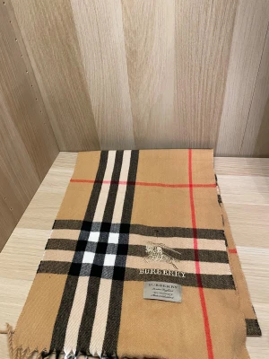 Rutig beige halsduk från Burberry - Snygg halsduk från Burberry i klassiskt rutigt mönster med beige bas och inslag av svart, vit och röd. Halsduken har fransar i ändarna och Burberrys logga broderad i guld. Perfekt accessoar för att lyfta din outfit med lyxig känsla.