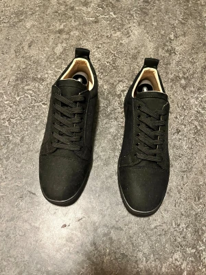 Louboutin Skor - Säljer ett par svarta sneakers från Louboutin med klassisk röd sula och snygg snörning. Skorna har rund tå, platt sula och är tillverkade i mocka med beige insida. Perfekt för dig som gillar stilrena och exklusiva detaljer.