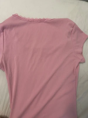 Rosa ribbad topp med spets och rosett - Supersöt ljusrosa topp med ribbad struktur, fyrkantig halsringning dekorerad med spetskant och en liten rosett framtill. Kortärmad och figurnära passform, perfekt för dig som gillar romantiska detaljer och mjuka färger.