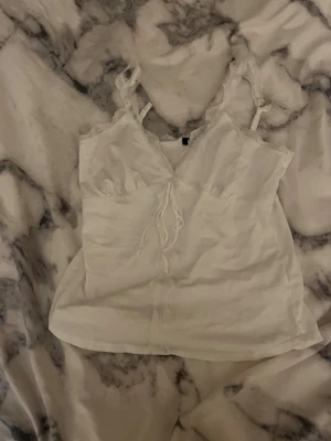 Vit spetsdetaljerad linnetopp DKNY - Supersöt vit linnetopp från DKNY med spetsdetaljer på axelbanden och runt halsringningen. Toppen har en v-ringad front med knyt och är tillverkad i mjuk bomull. Perfekt för sommarens alla tillfällen, enkel att styla med jeans eller kjol.