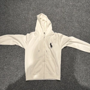 Vit Polo Ralph Lauren big pony  - Säljs då jag gått upp några pannor och den är för liten                                                                                 Snygg vit hoodie från Polo Ralph Lauren med stor svart broderad logga på bröstet och siffran 3 på ena ärmen. Tröjan har huva med snörning, dragkedja framtill och två fickor. Perfekt för dig som gillar klassisk streetstyle och vill ha något som sticker ut.