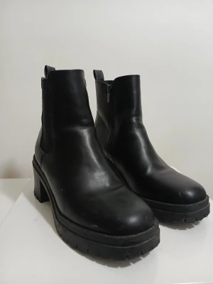 Svarta chunky boots med klack - Säljer ett par svarta boots med grov sula och blockklack. Skorna har rund tå, dragkedja på sidan och är tillverkade i skinnimitation. Stilrena och edgy, perfekta för dig som gillar chunky look och vill ha lite höjd.