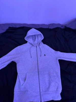 Grå hoodie från Polo Ralph Lauren - Snygg grå hoodie från Polo Ralph Lauren med dragkedja och huva med snörning. Klassisk logga broderad på bröstet och två stora fickor framtill. Perfekt för chill dagar och enkel att matcha med jeans eller joggers.