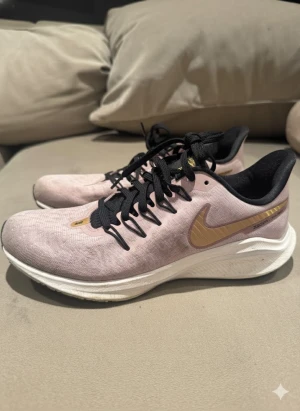 Nike Air Zoom Vomero 14  - Nike Air Zoom Vomero 14 i snygg rosa färg med guldfärgad swoosh. Skorna har svart snörning, vit platt sula och mesh-ovandel som andas. Perfekta för dig som gillar att springa eller vill ha en sportig look. Lätta och bekväma med responsiv dämpning. 