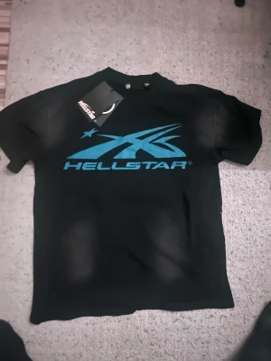 Svart Hellstar t-shirt med blått tryck - Svart t-shirt från Hellstar med stort blått grafiskt tryck på både fram- och baksidan. Framsidan har Hellstar-loggan och baksidan har en fet blå cirkel med en profil och stjärna. Klassisk rund hals och korta ärmar. Materialet är mjuk bomull.