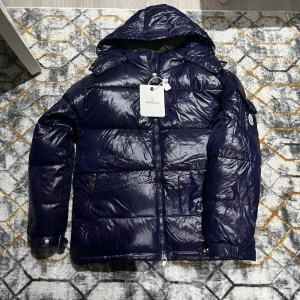 Mörkblå Moncler pufferjacka med huva - Snygg mörkblå pufferjacka från Moncler med glansig finish och huva. Jackan har dragkedja framtill, två fickor med dragkedja och klassisk Moncler-logga på ärmen. Perfekt för kalla dagar och riktigt stilren look.