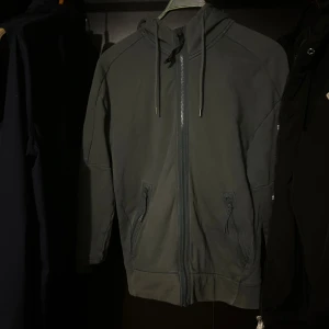 Grå hoodiejacka med goggles från C.P. Company - Säljer en grå hoodiejacka från C.P. Company med ikoniska goggles på huvan. Jackan har dragkedja framtill, två fickor med dragkedja och justerbar huva med snörning. Perfekt för dig som gillar streetwear och vill sticka ut med en unik detalj.