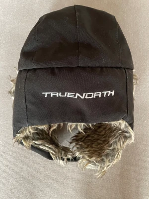 Svart pälsfodrad trappermössa True North - Säljer en svart trappermössa från True North med mjukt, beigegrått fuskpälsfoder. Mössan har öronlappar med knäppning och broderad logga på sidan. Perfekt för kalla vinterdagar när du vill hålla dig varm och ändå se chill ut.