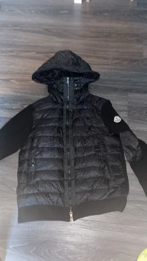 Svart Moncler pufferjacka XL - Svart pufferjacka från Moncler med huva och stickade ärmar. Jackan har quiltad front, dragkedja och Moncler-logga på ärmen. Materialmix med polyester och stickad textil ger en cool kontrast. Perfekt för dig som gillar streetstyle och exklusiva märken.