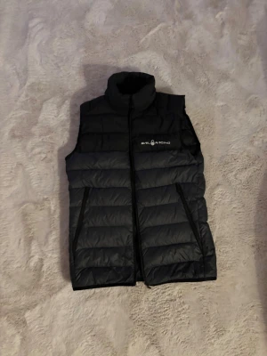 Svart dunväst från Sail Racing - Svart dunväst från Sail Racing med hög krage och horisontella sömmar. Västen har dragkedja framtill, två snedställda fickor med dragkedja och logga på bröstet. Perfekt för lager-på-lager och stilren look.