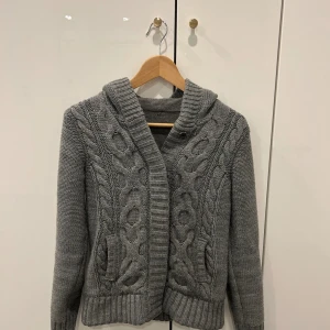 Grå stickad cardigan med huva - Mysig grå stickad kofta med huva och grovt flätmönster framtill. Koftan har ribbade muddar och bred krage, samt två fickor på sidorna. Perfekt för kyliga dagar och enkel att matcha med det mesta.