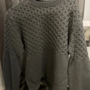 Grå stickad tröja med strukturmönster - Säljer en grå stickad tröja med snyggt strukturmönster över bröst och ärmar. Tröjan har rund hals och långa ärmar med ribbade muddar. Perfekt för dig som gillar stilrena och mysiga plagg med lite extra detalj på ovansidan.