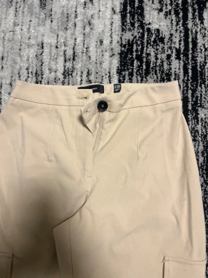 Beiga cargobyxor från Vero Moda - Stilrena beiga cargobyxor från Vero Moda med raka ben och stora fickor på sidorna. Byxorna har hög midja, knapp och dragkedja framtill. Perfekta för en avslappnad och trendig look.