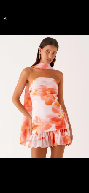 Blommig kort klänning med volang - Säljer en superfin axelbandslös kort klänning med blommigt mönster i rosa, orange och vitt. Klänningen har volangkant nedtill och en matchande scarf som hänger bak. Perfekt för sommarens alla tillfällen, riktigt söt och trendig look. Prislappen är på 